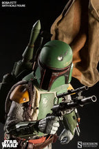Close-up of the Boba Fett sixth-scale figure holding a blaster rifle, with a green jetpack, chipped helmet, yellow shoulder armor, and brown cape behind him