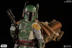 Boba Fett sixth-scale figure holding a blaster rifle, wearing chipped green armor, yellow shoulder plates, a brown cape, and a jetpack on a black background.